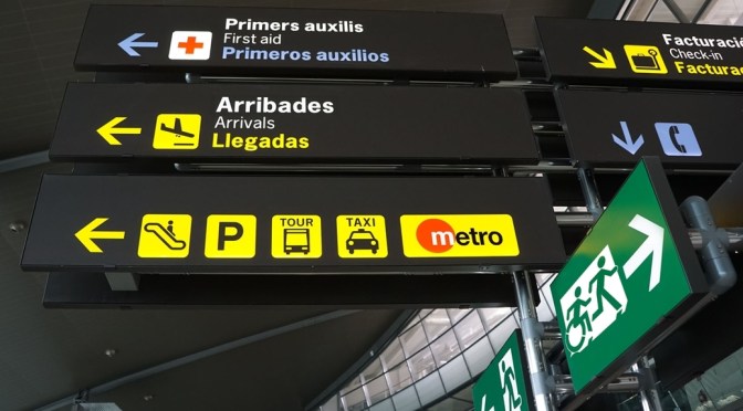 Airport signage board stating "Metro" with an exit sign pointing to the right with a person running followed by a person using a wheelchair, moving in the same style as the running person. On the left of the signage board is another exit sign, but this one only has the running person moving to the left. The sign shows that an accessible exit path is to the right, whilst the non-accessible exit path is to the left.