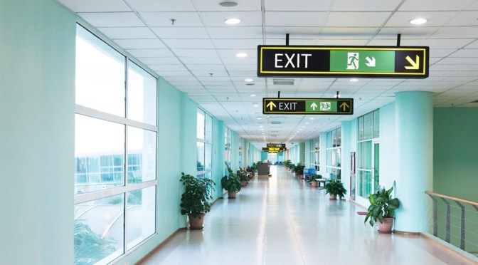 Universal Design Meets the Exit Sign Accessible Means of Egress Icon at hospital. Image shows two different green exit signs. The first has a person running to the right with an arrow pointing to the stairs, the second has the running man followed by a person using a wheelchair, moving in the same style as the running person, they are moving the left through a doorway.
